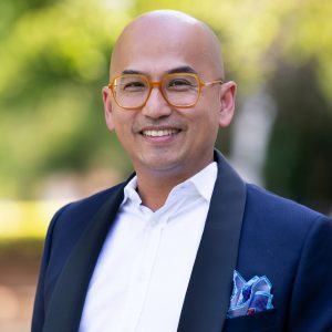 A headshot of a man against a green outdoor background.